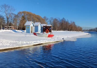 Talviuinti Oulussa. Tuiran talviuintipaikassa lumen peittämä ranta ja vaaleansiniset uimakopit. Sininen vesi lainehtii vapaana.